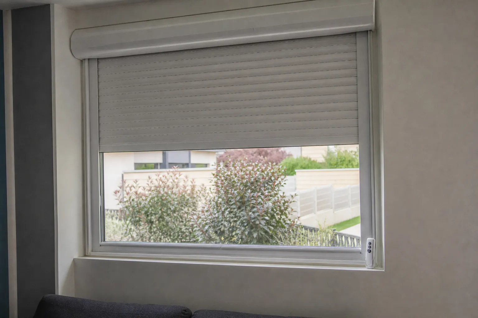 Volet roulant aluminium blanc à mi-hauteur sur une fenêtre, télécommande posée sur le rebord intérieur